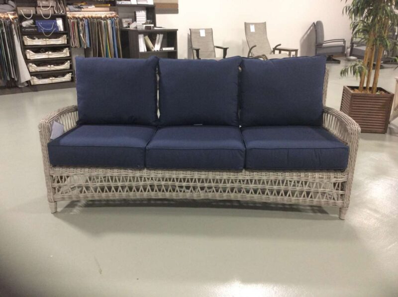 A wicker three-seater sofa with navy blue cushions in a showroom setting, surrounded by various furniture items including a fireplace and decoration pieces.