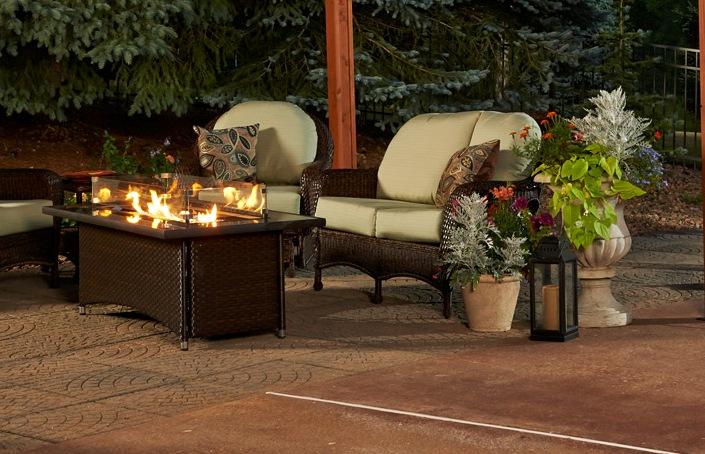 An outdoor patio setup at dusk, featuring a dark wicker furniture set with cushions, a lit stove in the center, and vibrant floral arrangements beside the seating.