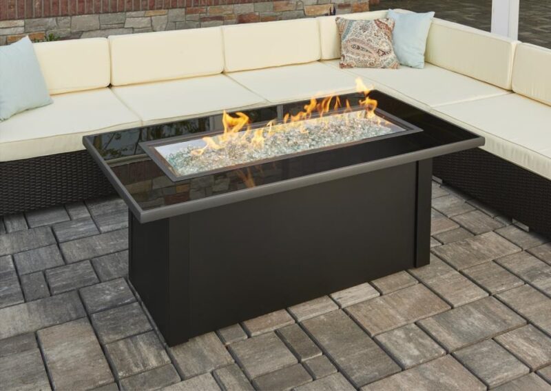 A modern outdoor gas fire pit with bright flames, surrounded by a black metal frame on a stone-paved patio, complemented by a cream cushioned corner sofa.