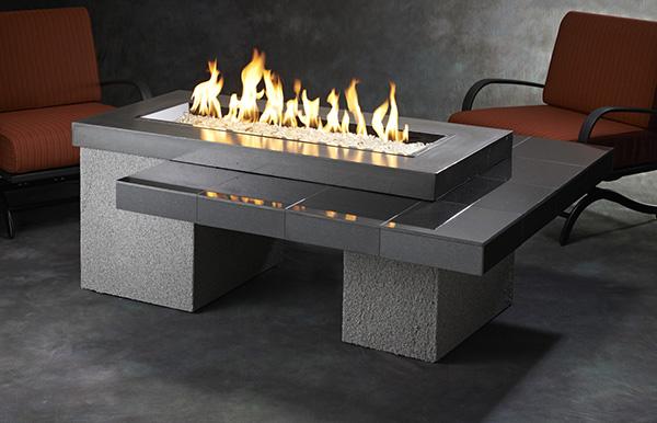 A modern outdoor fire pit table with flames burning in the center, set on a textured grey base with a sleek black tabletop. Two red chairs are placed nearby on a concrete floor.