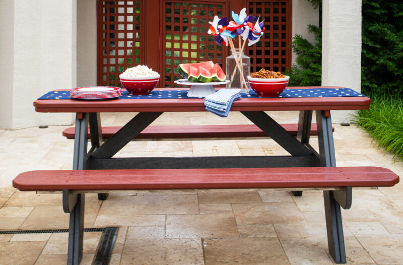 A picnic table decorated with a patriotic theme, featuring red, white, and blue tablecloth, plates, and pinwheels, with bowls of popcorn and pretzels, slices of watermelon,
