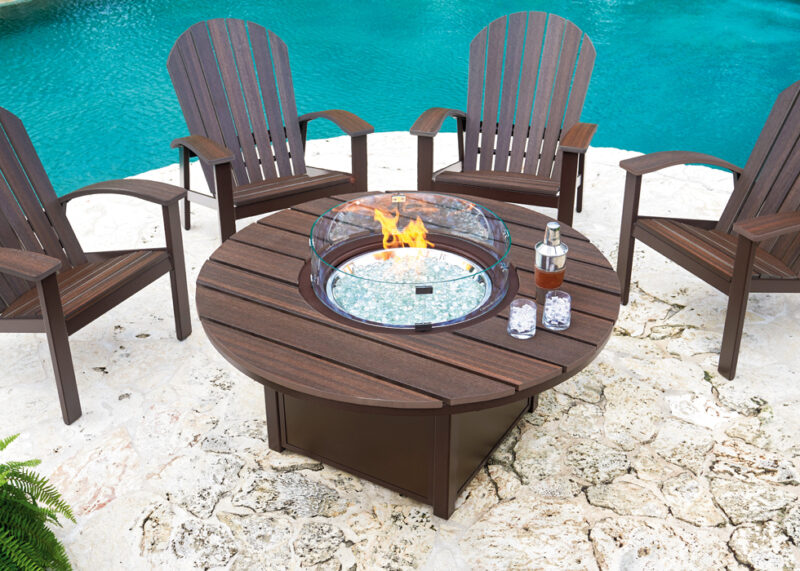 An outdoor patio setup with a circular, wooden fire pit insert table surrounded by four dark brown adirondack chairs, on a white pebbly surface near a clear blue swimming pool.