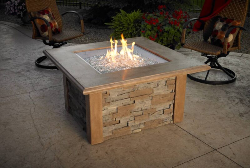 A square fire pit with a lit flame, surrounded by glass beads, is set on a patio. Nearby are two chairs with patterned cushions, and a flowerbed with red blooms enhances the cozy outdoor