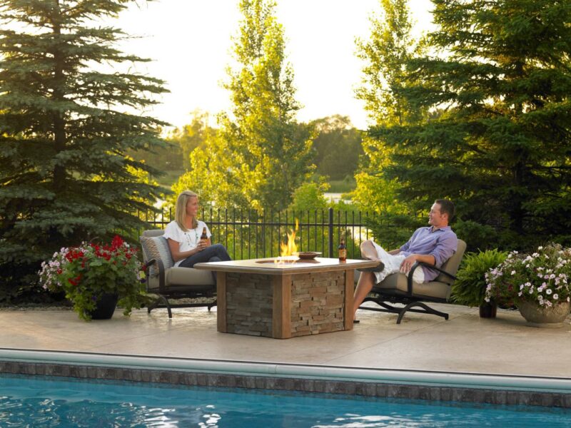 A man and a woman relax on patio chairs by a pool at sunset, surrounded by lush greenery and a stone fire pit insert table between them.