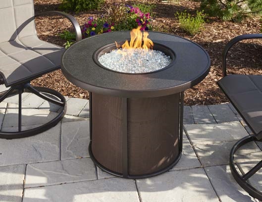 A modern outdoor fire pit insert with a circular design and a blue flame, surrounded by four curved metal chairs on a paved patio area.