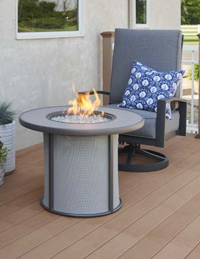 A cozy outdoor setting featuring a modern patio chair with a grey cushion and a decorative blue pillow next to a circular fireplace table with flames, on a wooden deck.
