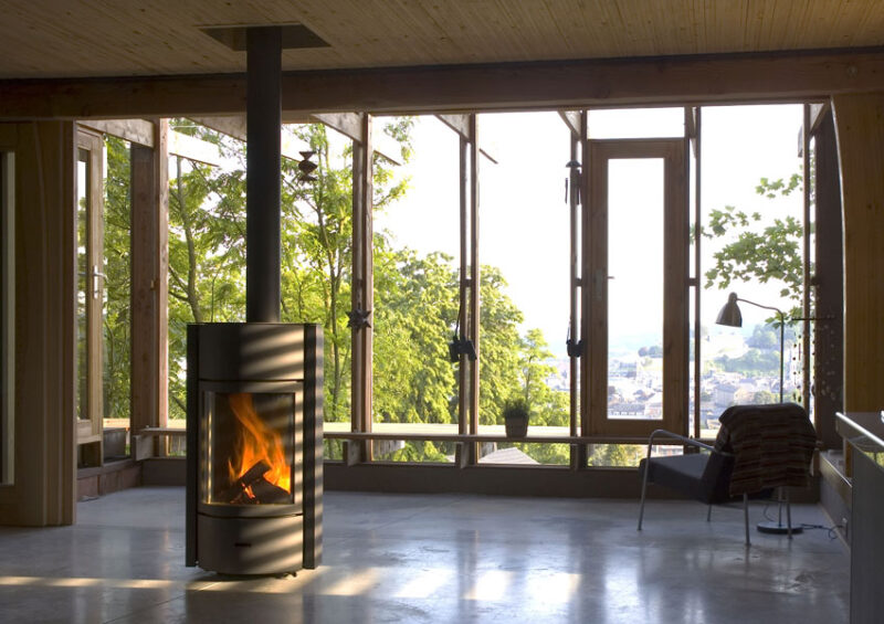 A modern, spacious living room with large windows overlooking a scenic view, featuring a freestanding fireplace insert with visible fire, wooden walls and flooring, and minimalist furniture.