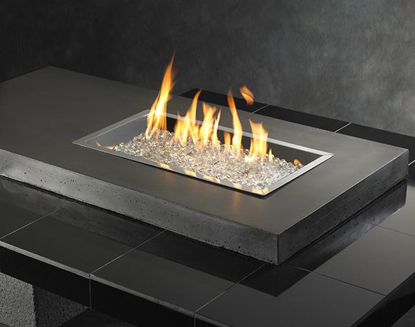 Modern rectangular tabletop fireplace burning on a gray concrete surface, surrounded by crushed glass, in a minimalist setting.