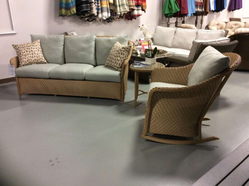 A furniture display featuring a light beige sofa with patterned cushions, two matching wicker chairs, a rocking chair, and a small wood coffee table, set in a well-lit store near a cozy