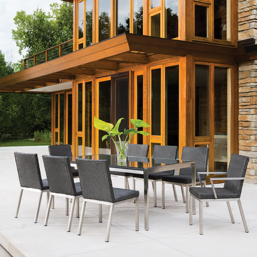 Outdoor dining area with a modern table and chairs set on a patio, in front of a contemporary house with large glass windows, wooden details, and an elegant fireplace.