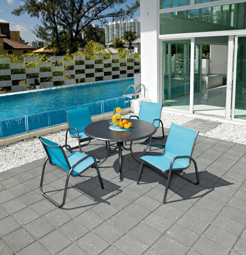 Outdoor patio setting with a round table and four turquoise chairs, a bowl of oranges on the table, adjacent to a swimming pool with a tiled fountain wall, and an integrated fire pit, under a clear
