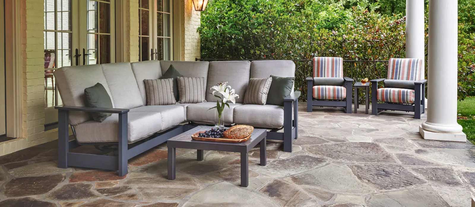 Outdoor patio with a modern sectional sofa offset by two striped chairs, centered around a low coffee table, set on a stone floor with lush greenery in the background and warmed by an elegant fireplace insert.