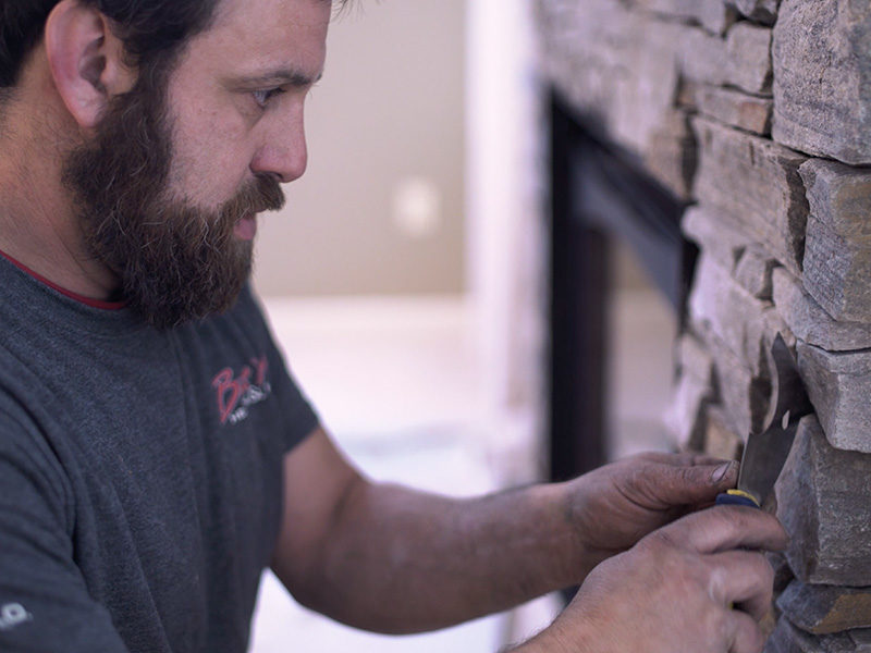 A man with a beard is carefully applying mortar to insert a fire pit into the stone wall, focusing intently on his work.