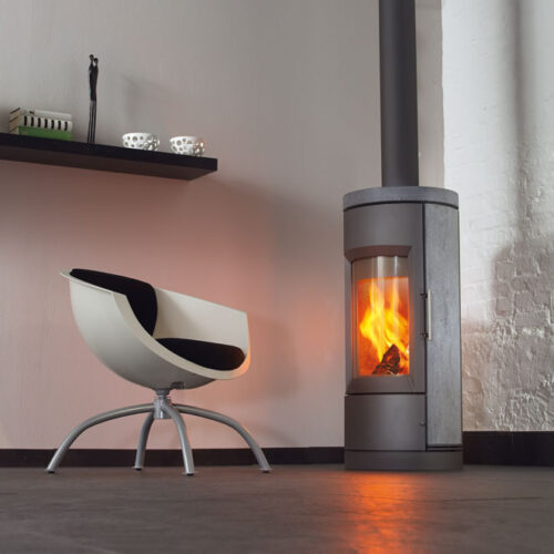 A modern room featuring a freestanding metal fireplace with a visible fire, beside a stylish black and white chair, set against a white brick wall with decorative items on a shelf.