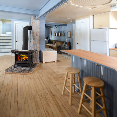 A cozy, well-lit kitchen with wooden flooring and blue accents. features include a central stove, a dining table, bar stools, and modern appliances like a fridge.