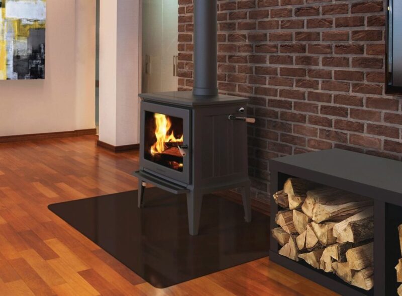 A modern wood-burning stove with visible flames is set against a brick wall in a cozy room. a wood storage box sits nearby on a polished wooden floor.
