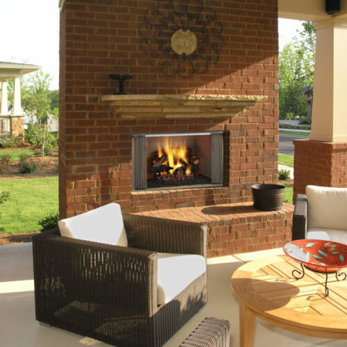 A cozy outdoor patio featuring a brick fireplace, black wicker chairs with white cushions, a round wooden table, and a decorative red metal tray, set against a backdrop of lush greenery and suburban homes.
