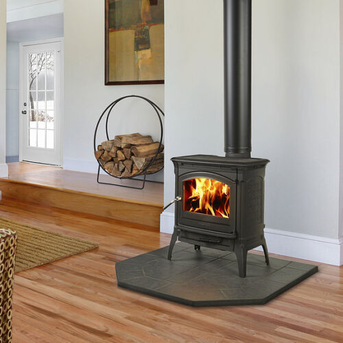 A cozy living room with a lit wood-burning stove on a gray tile platform, surrounded by hardwood floors. a sofa, wood log holder, and stairway are visible in the background.