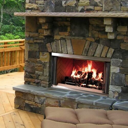 An outdoor stone fireplace with a roaring fire, set on a wooden deck bordered by a lush green forest. a cozy beige sofa is in the foreground.