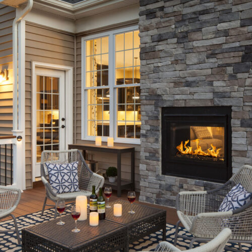 Cozy outdoor patio with a stone fireplace, two wicker chairs with cushions, and a warm glow from the house windows during twilight.