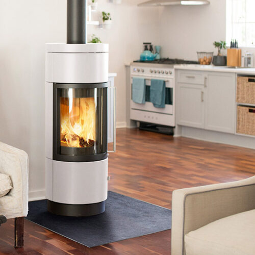 A modern, cylindrical, white wood-burning stove with a large glass door, situated in a cozy living room with light wood floors and a blue mat.