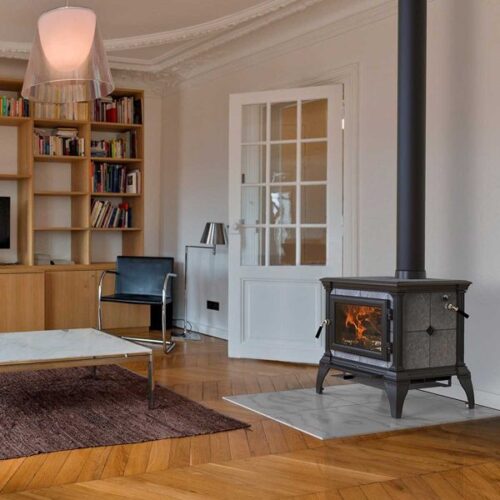 A cozy room with a lit wood stove, a modern black chair, a wooden desk, and a large rug on a hardwood floor. white walls and elegant moldings enhance the warm, inviting atmosphere.