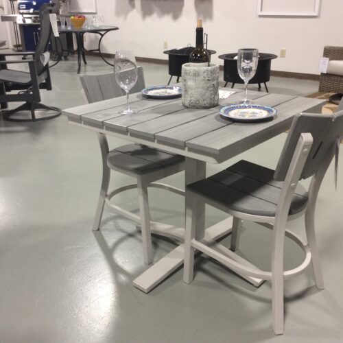 A modern dining table set with grey and white finishes, featuring two chairs and a bench, elegantly set with plates and wine glasses, displayed in a furniture showroom.