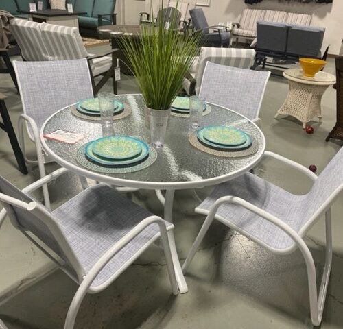 A Gardenella patio furniture display featuring a round glass table and four white chairs with blue seat cushions in an indoor showroom. Green plants and other furniture pieces are visible in the background.