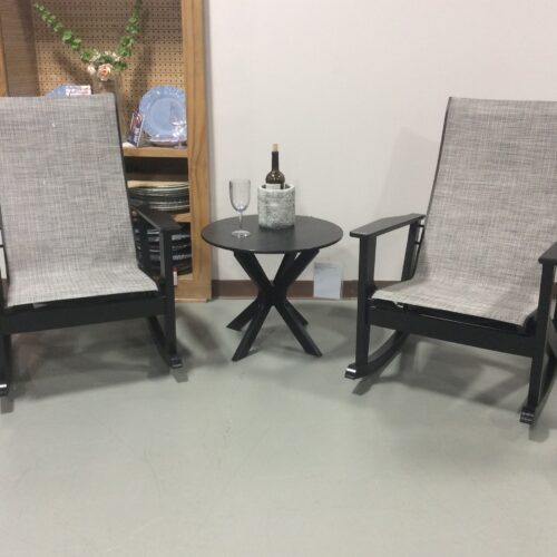 Two black outdoor chairs with gray mesh fabric facing a small round table holding a wine bottle and two glasses, set against a white wall with decor shelves.