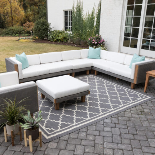 Elegant outdoor patio furniture arrangement featuring a light gray Catalina sectional sofa, matching chairs and ottoman, set on a textured rug, with a wooden coffee table, all surrounded by a lush garden.