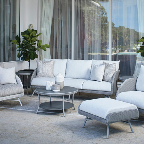 Outdoor patio exuding the essence of modernity with wicker furniture featuring white cushions, including a sofa, chairs, and a chaise lounge, arranged around a round coffee table near sleek glass doors.