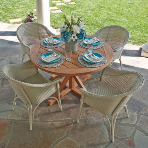 Outdoor dining setup with a round wooden table and four wicker chairs on a stone patio. the table is set with plates, cutlery, and a vase with blue flowers.