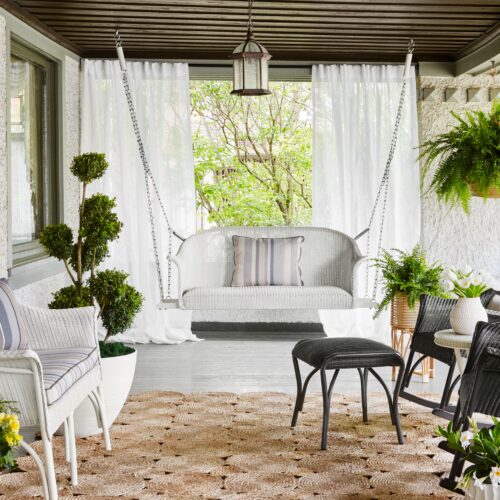 A cozy, well-decorated porch with a hanging bench, chairs, lush plants, curtains, and a textured rug. the space exudes a relaxing atmosphere with a blend of modern and rustic elements.