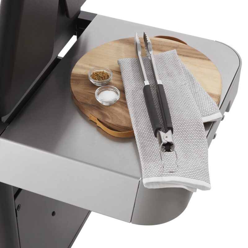 A Weber Genesis S-435 stainless steel grill with a side tray holding a round wooden cutting board, a small bowl of spices, and tongs resting on a folded metallic kitchen towel.