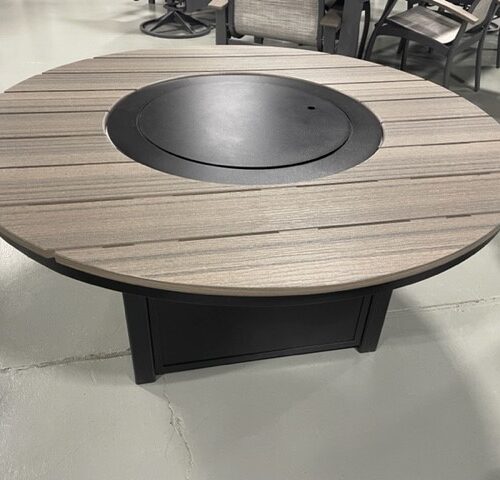 Round wooden table with a central black metal fire pit, surrounded by Newport Adirondack chairs, displayed on a grey concrete floor in a showroom setting.