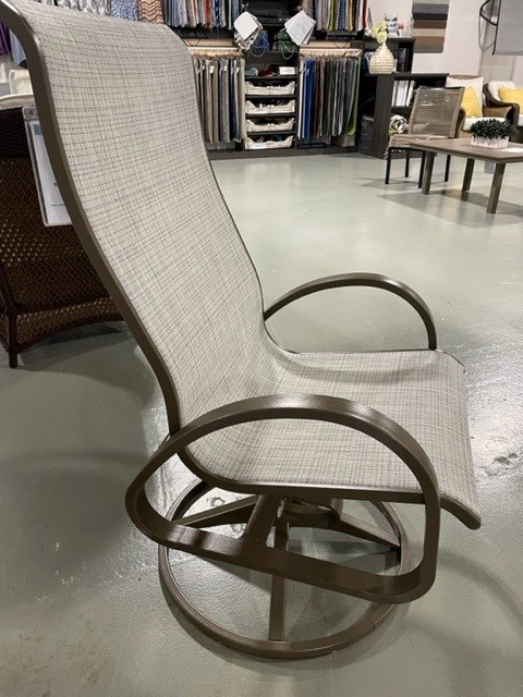 A modern Aruba II rocking chair with a unique circular base and a woven tan upholstered back and seat, situated on a polished concrete floor in an interior setting, surrounded by various textiles.