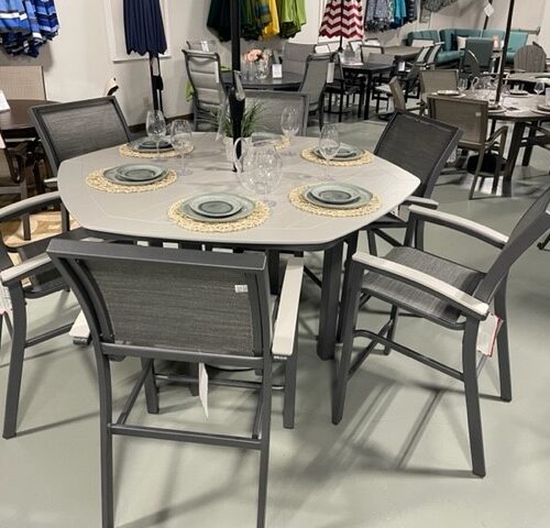 An outdoor dining set featuring a round, white Bazza tabletop and four dark gray chairs with armrests, displayed in a store with various other furniture in the background.