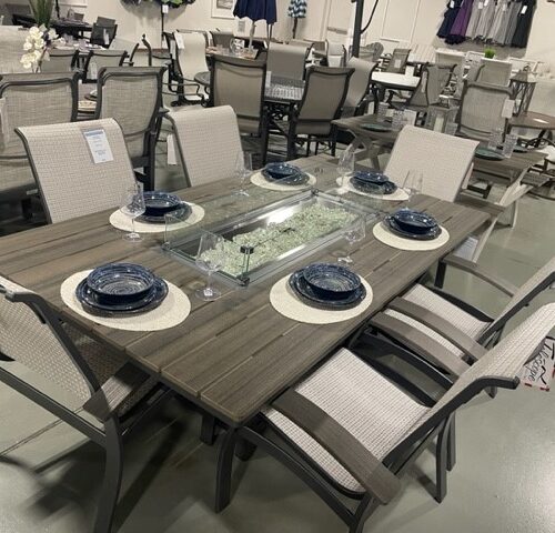A modern Belle Isle Sling outdoor dining set featuring a rectangular table with a glass center and six chairs, displayed in a furniture showroom. The table is set with blue dishes and decorative plants.