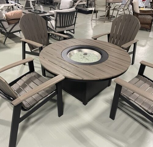 A round outdoor table with a central fire pit and four Newport Adirondack chairs, displayed on gray flooring in a showroom setting. The furniture features dark frames and wood accents.
