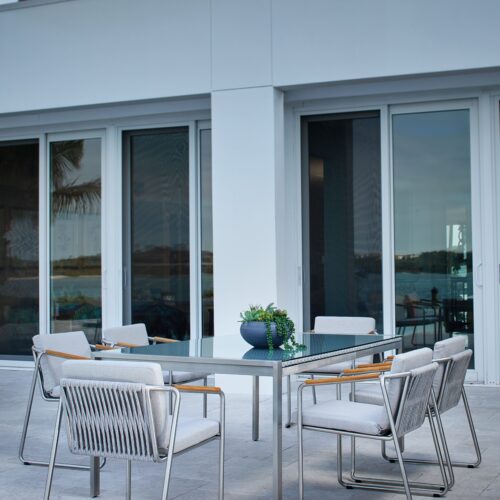 A modern outdoor dining area with four stylish metal chairs around a wooden table, set against a contemporary white home with large glass doors and palm trees in the background.