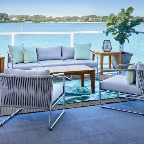 A modern outdoor patio setting with a sofa and armchairs around a wooden coffee table. the furniture is positioned on a balcony overlooking a tranquil blue river. decor includes cushions and potted plants.