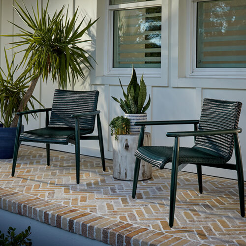 Two black wicker chairs on a brick-patterned patio with a small table between them, near a potted plant and under the window of a Fairview house.