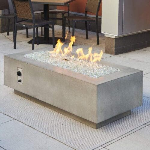 A rectangular, modern outdoor gas fire pit with a concrete finish, adorned with blue glass crystals, flames visible, set on a patio with chairs in the background.