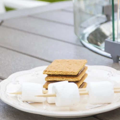 A plate with marshmallows and a s'more sits on a table next to the Fire & Ice Table by Breezesta, with outdoor chairs and cushions in the background, suggesting a cozy