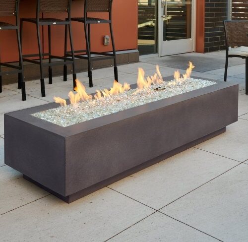 A modern outdoor rectangular fire pit with a blazing flame, surrounded by glass pebbles, situated on a patio area with seating in the background.