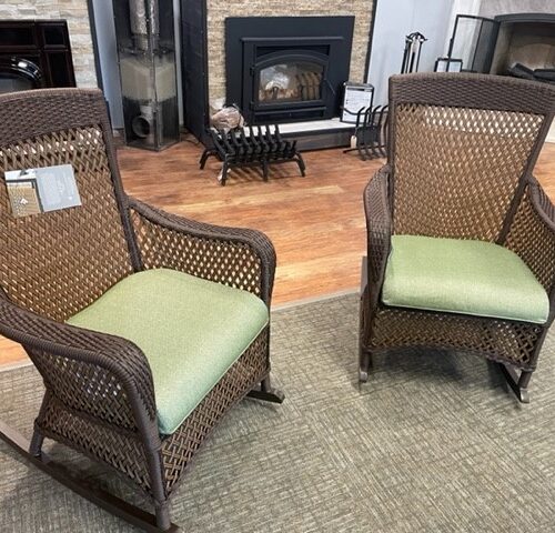 Two Grand Traverse wicker rocking chairs with green cushions displayed in a showroom with various fireplace models in the background.