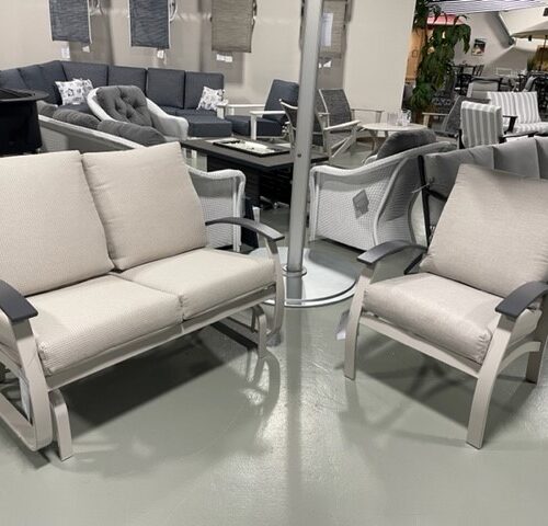 A display of Belle Isle Cushion patio furniture featuring a loveseat and two chairs with light gray cushions, arranged around a small round table with a central pole, in a brightly lit store.