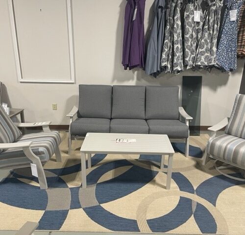 A cozy lounge area in a clothing store on St. Catherine Street features a grey sofa, two striped armchairs, and a white coffee table on a blue circular pattern rug, surrounded by clothing racks