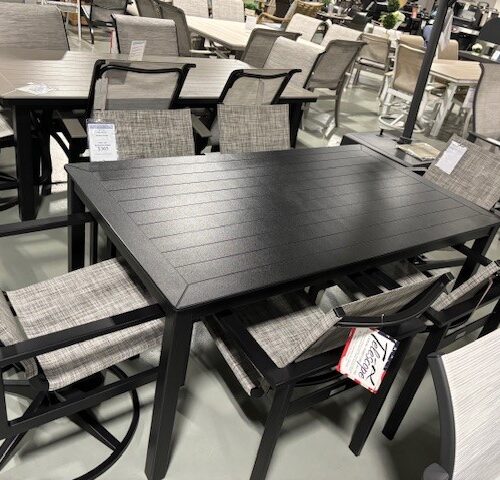 A display of Tribeca outdoor furniture featuring dark-framed tables and chairs with gray woven seat and backrest, set up in a store showroom with other furniture items in the background.