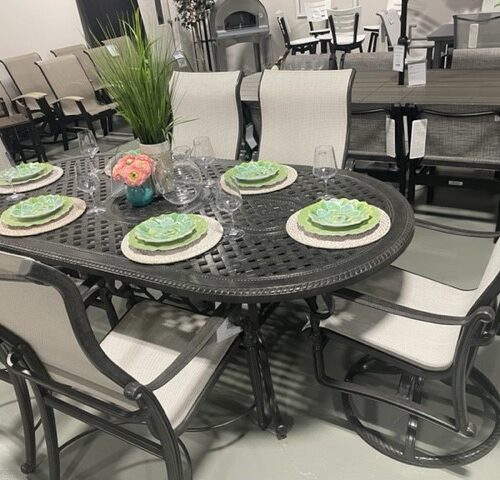 A display of Gensun Bel Air outdoor dining furniture in a showroom, featuring a dark rattan table set with white and green place settings and surrounded by coordinating chairs.